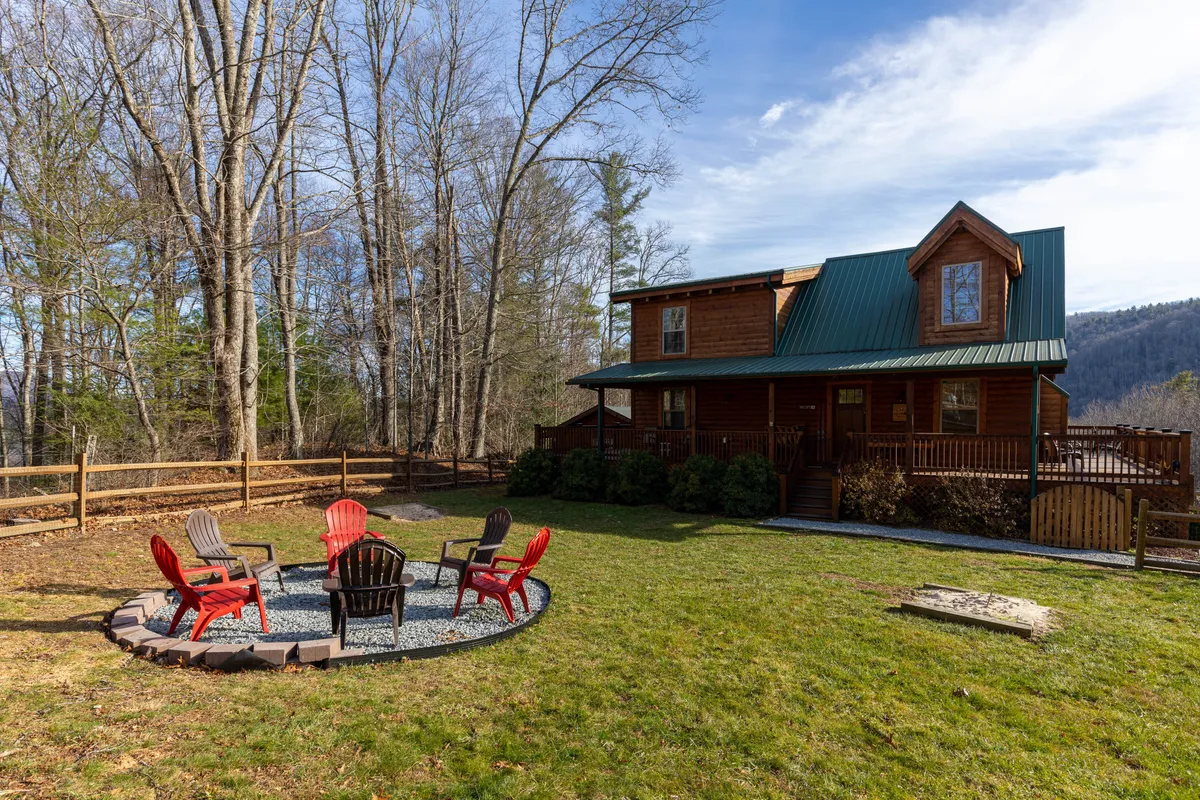 An inviting view of the fire pit area nestled among trees, with a wooden house in the background. The seating around the fire pit and the greenery emphasize outdoor relaxation, communal experiences, and the natural setting of the property.