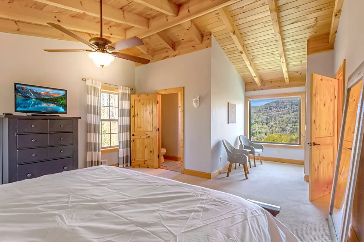 A serene bedroom with a plush bed, accent chairs, generous window, TV, and wooden doors, awash in natural light. The scene is calm and inviting, suggesting a restful retreat with modern comforts.