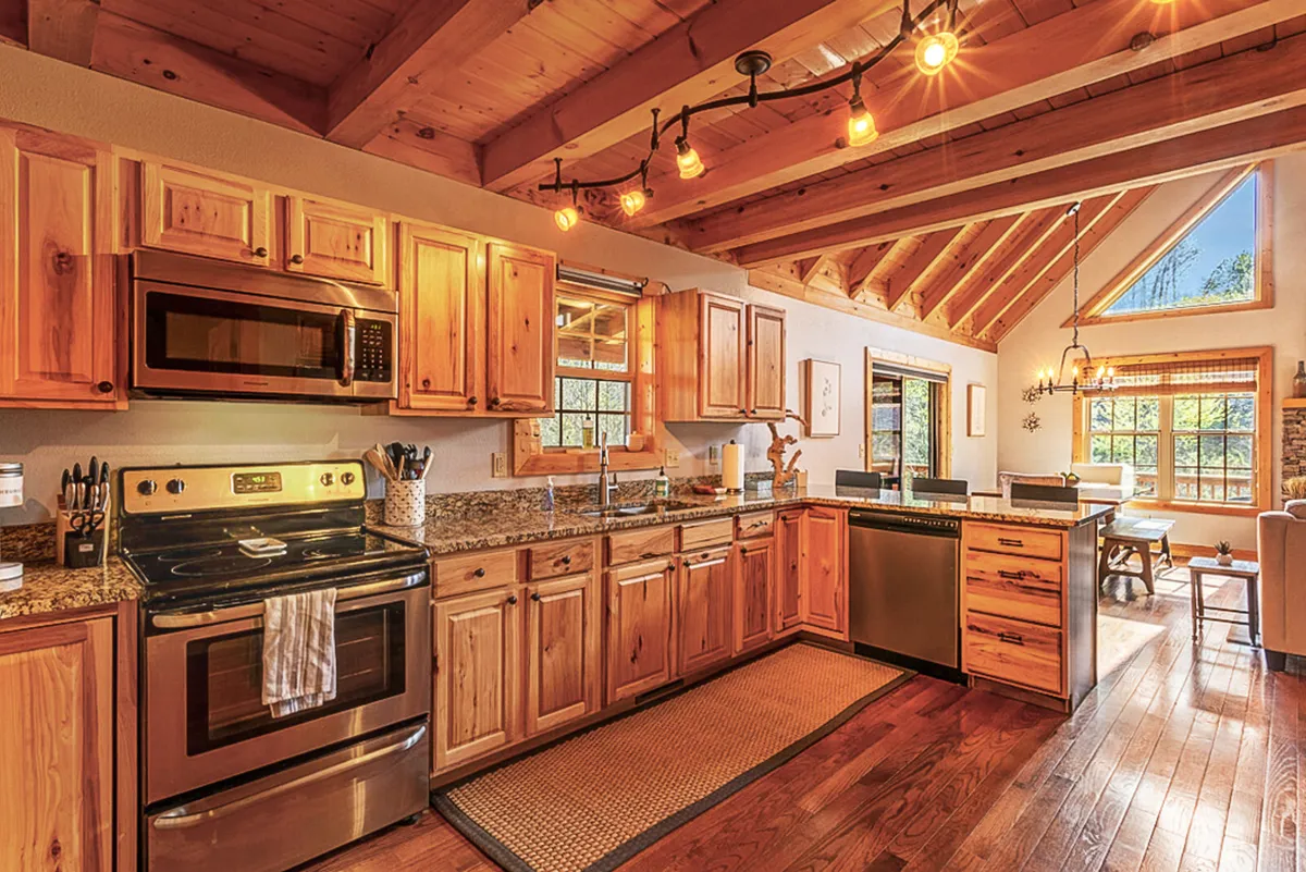 A bright kitchen featuring granite countertops, stainless steel appliances, wooden cabinetry, open shelving, and a large window. The island offers additional workspace and an informal spot for quick bites or conversation during meal prep.