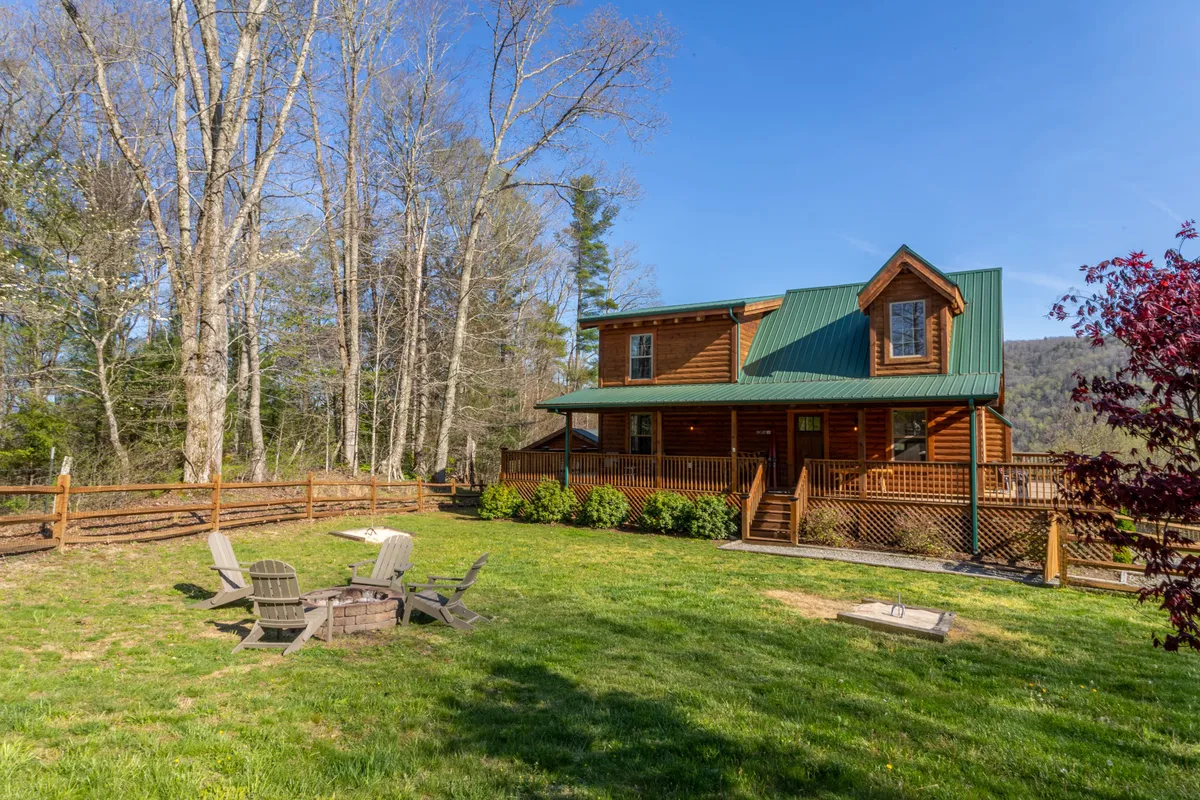 A cozy log cabin exterior with a green metal roof, surrounded by a beautifully landscaped garden. A fire pit with Adirondack chairs invites guests to gather outdoors, and the mountain backdrop completes the tranquil, family-friendly setting.