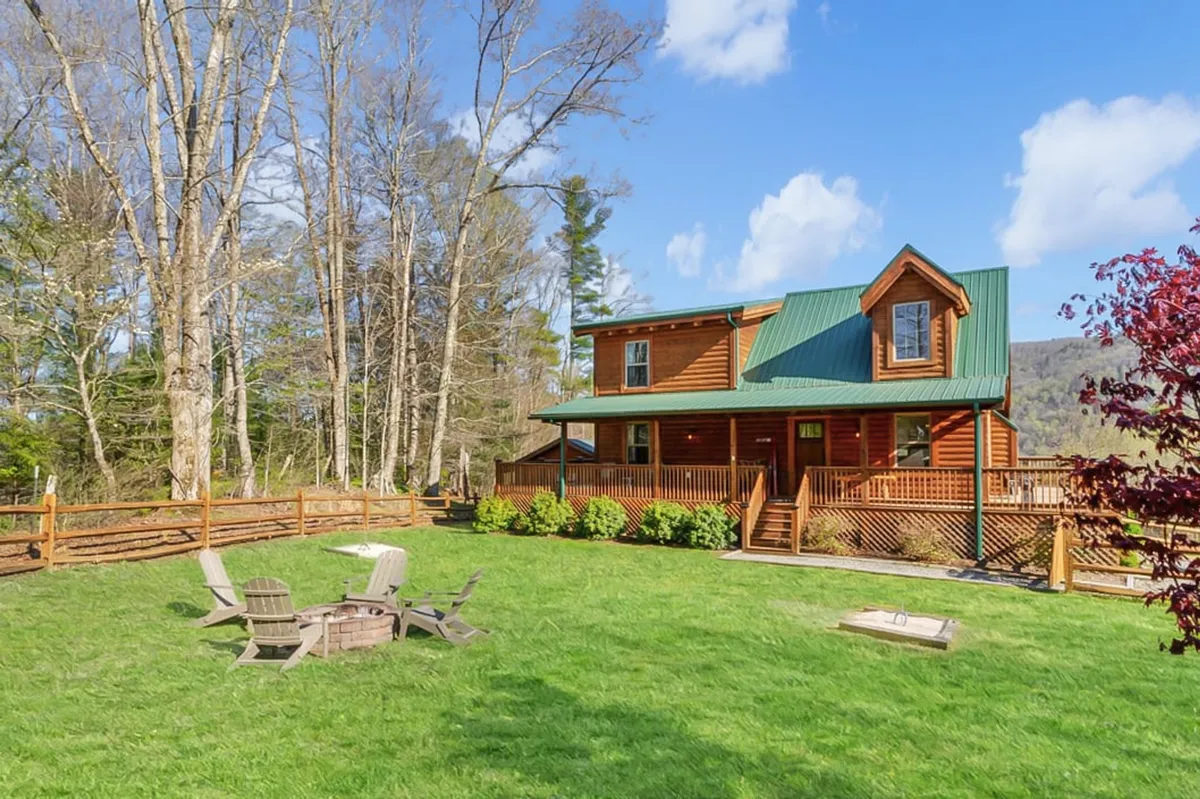 A broad exterior view of the property, showing the house surrounded by trees, a fire pit area with seating, and a wooden deck. The tranquil yard space is partially visible, suggesting a spacious and welcoming environment ideal for outdoor activities and pet play.