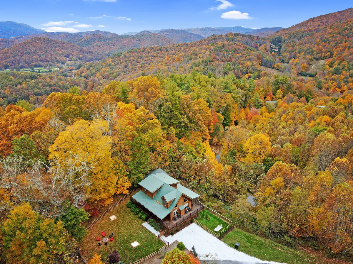 The image features a charming log cabin nestled among vibrant autumn trees, with expansive mountain vistas in the background. An inviting outdoor seating area is visible on the porch, perfectly positioned for sunset views and relaxation. The scene evokes serenity and the promise of outdoor adventures, hinting at opportunities for hiking, family gatherings, and evenings by the fire pit under the stars.