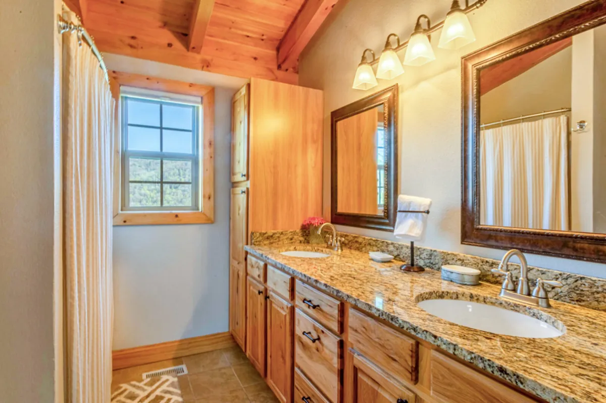 A bright, spa-inspired bathroom with a double vanity, large mirrors, wood cabinetry, and a window with a view. The space feels spacious, clean, and ideal for both morning routines and winding down.